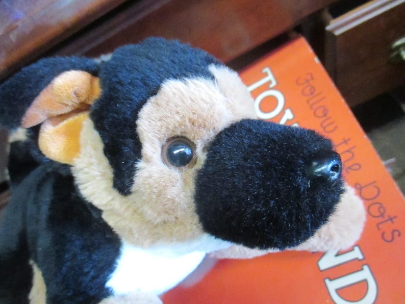 May include: A plush German Shepherd dog toy with black, tan, and cream-colored fur. The toy has a black nose and a detailed face. The toy is resting on an orange book with the words "Follow the Dots" visible.