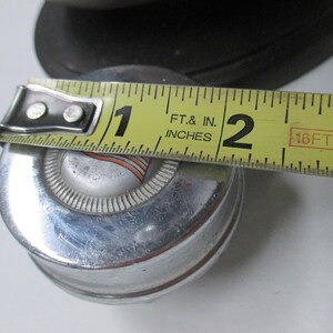May include: A circular chrome hubcap with a central silver emblem featuring orange and black stripes. A yellow measuring tape is placed across the top, indicating a diameter of approximately 2.5 inches for the center portion of the object.