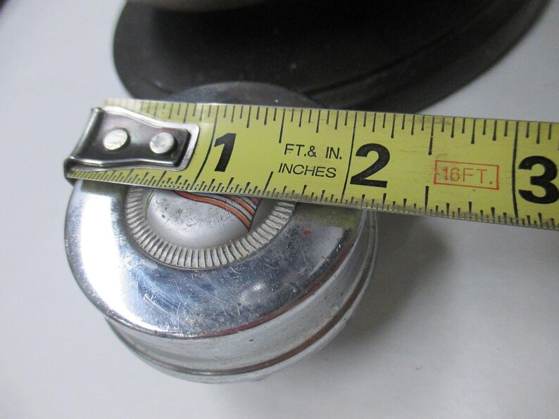 May include: A circular chrome hubcap with a central silver emblem featuring orange and black stripes. A yellow measuring tape is placed across the top, indicating a diameter of approximately 2.5 inches for the center portion of the object.