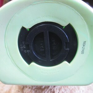 May include: Close-up of the bottom of a light green piggy bank. The black coin slot is in the center, with the word "OPEN" visible. The text "Monogram International Inc." and "YA34-21H20" are also printed on the bank.