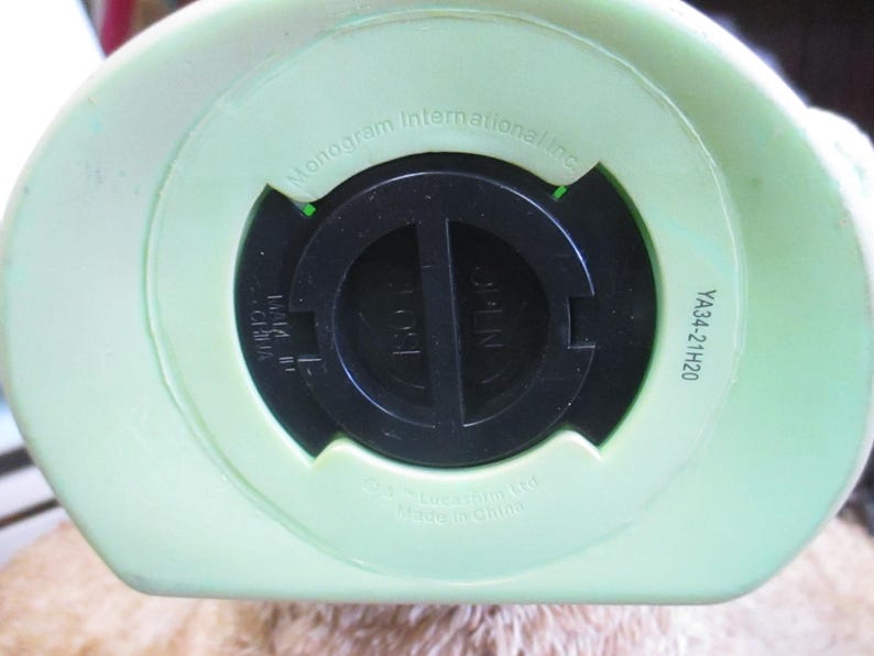 May include: Close-up of the bottom of a light green piggy bank. The black coin slot is in the center, with the word "OPEN" visible. The text "Monogram International Inc." and "YA34-21H20" are also printed on the bank.
