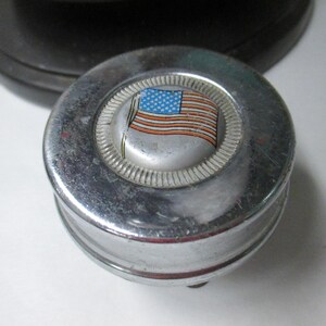 May include: A circular chrome metal object with a raised center featuring a graphic of the American flag. The metallic surface has a polished finish with a textured ring surrounding the central flag emblem, showing signs of wear and light scratches.
