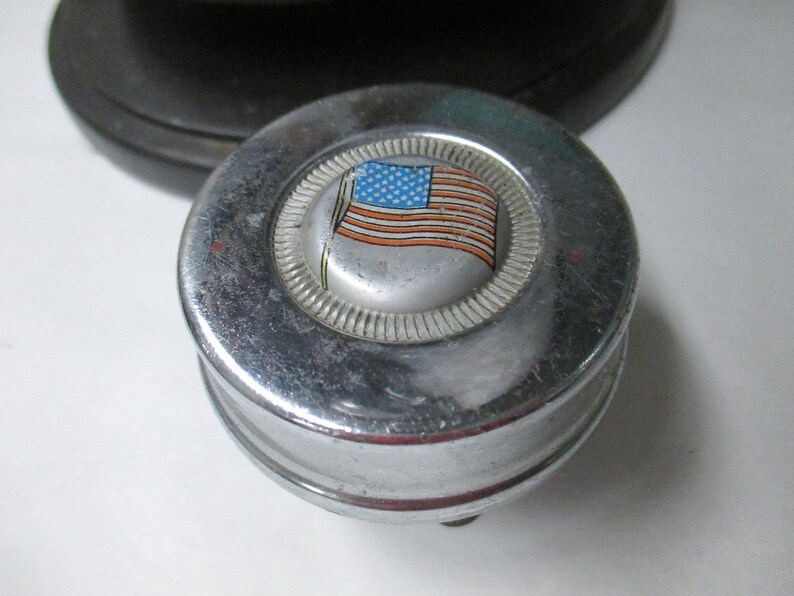 May include: A circular chrome metal object with a raised center featuring a graphic of the American flag. The metallic surface has a polished finish with a textured ring surrounding the central flag emblem, showing signs of wear and light scratches.