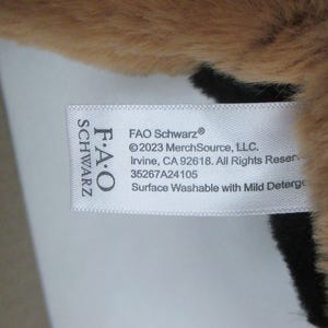 May include: Close-up of a plush toy with brown and black fur. A white tag displays the FAO Schwarz logo and product information, including the text "Surface Washable with Mild Deterge."