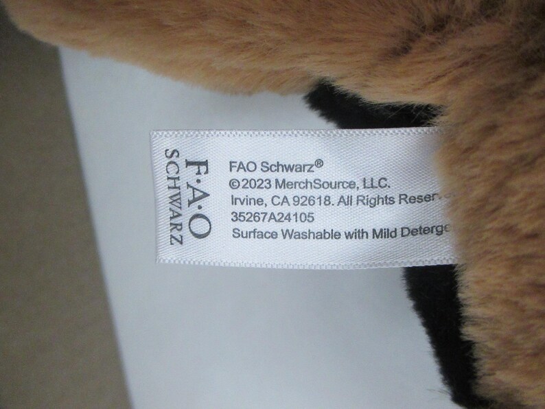 May include: Close-up of a plush toy with brown and black fur. A white tag displays the FAO Schwarz logo and product information, including the text "Surface Washable with Mild Deterge."
