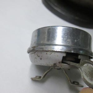 May include: A vintage bicycle bell with a polished chrome dome and a weathered white base. The bell features a side-mounted metal lever for activation and two small metal mounting brackets extending from the bottom of the circular housing.