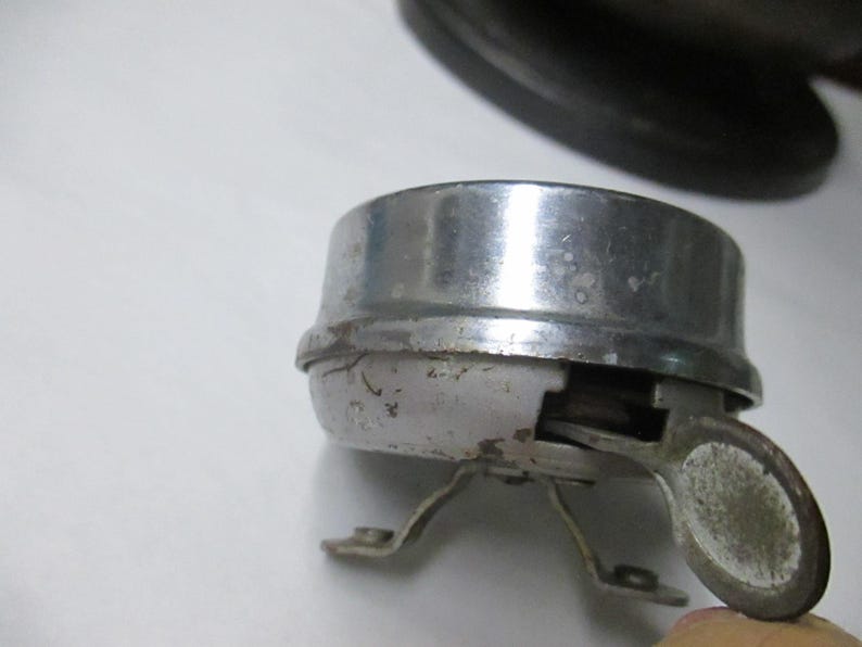 May include: A vintage bicycle bell with a polished chrome dome and a weathered white base. The bell features a side-mounted metal lever for activation and two small metal mounting brackets extending from the bottom of the circular housing.
