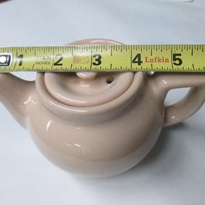 May include: A small, light tan ceramic teapot with a lid and handle. The teapot is approximately 5 inches long, as measured by a yellow tape measure. The teapot has a spout and a small handle for easy pouring.