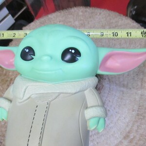 May include: A green figure with large black eyes and pink-lined ears, wearing a tan coat with a white collar. The figure is approximately 11 inches long, resembling a character from a popular science fiction series.