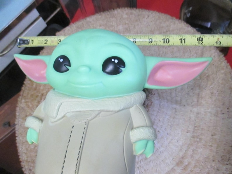 May include: A green figure with large black eyes and pink-lined ears, wearing a tan coat with a white collar. The figure is approximately 11 inches long, resembling a character from a popular science fiction series.