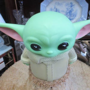 May include: A green figurine of a character with large ears and black eyes. The figurine is wearing a tan coat with a furry collar. The figurine is sitting on a tan, textured surface.