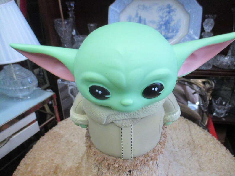 May include: A green figurine of a character with large ears and black eyes. The figurine is wearing a tan coat with a furry collar. The figurine is sitting on a tan, textured surface.