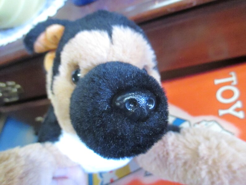 May include: Close-up of a plush toy dog with a black nose and black and tan fur. The toy has a white patch on its chest and is set against a blurred background. The word "TOY" is visible in the background.