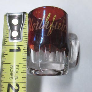May include: Clear glass shot glass with a handle, measuring approximately 1 inch tall. The top portion is a deep red color with the word "Northfield" in gold script. A yellow measuring tape is in the frame.