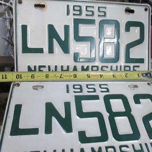 May include: Two vintage 1955 New Hampshire license plates. The plates are white with dark green lettering and numbers. The plates read "LN 582". A yellow measuring tape is placed across the plates, measuring approximately 12 inches.