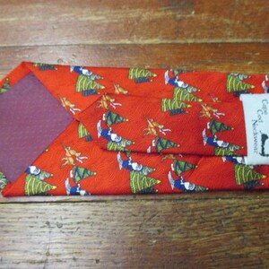May include: Red necktie with a repeating pattern of Christmas trees, figures, and reindeer. The tie has a burgundy backing and a label that reads "Cape Cod Neckwear".