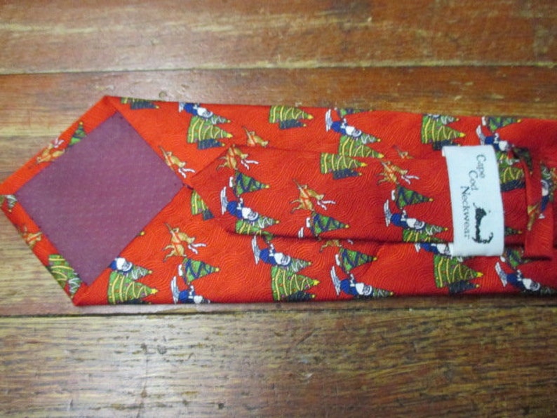 May include: Red necktie with a repeating pattern of Christmas trees, figures, and reindeer. The tie has a burgundy backing and a label that reads "Cape Cod Neckwear".