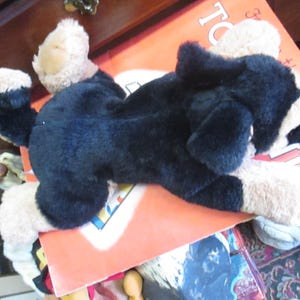 May include: A plush dog toy in black and tan, lying on an orange book. The toy has floppy ears and a soft, furry texture. The dog is lying down, with its paws extended. The toy is a classic children's item.