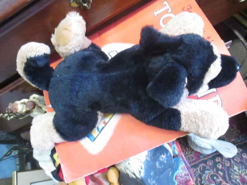 May include: A plush dog toy in black and tan, lying on an orange book. The toy has floppy ears and a soft, furry texture. The dog is lying down, with its paws extended. The toy is a classic children's item.