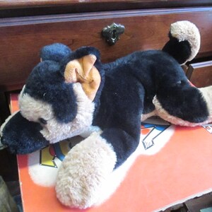 May include: A plush German Shepherd dog toy in black and tan, lying on a book. The toy has a soft, furry texture and is positioned on a book with visible text. The dog's ears are tan, and its paws are light brown.