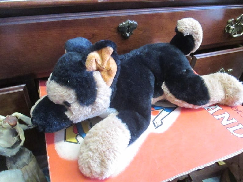 May include: A plush German Shepherd dog toy in black and tan, lying on a book. The toy has a soft, furry texture and is positioned on a book with visible text. The dog's ears are tan, and its paws are light brown.