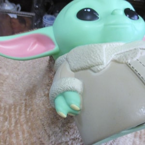 May include: A light green figurine of a character with large black eyes and pointed ears with pink insides. The figure is wearing a cream-colored coat with a white, furry collar and cuffs. The figurine is a popular character from a science fiction franchise.