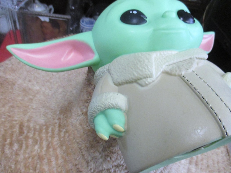 May include: A light green figurine of a character with large black eyes and pointed ears with pink insides. The figure is wearing a cream-colored coat with a white, furry collar and cuffs. The figurine is a popular character from a science fiction franchise.