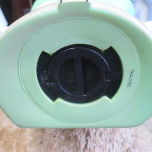 May include: Bottom view of a light green piggy bank with a black coin slot cover. The piggy bank has a round shape with a flat bottom. The text "YA34-21H20" is printed on the side.
