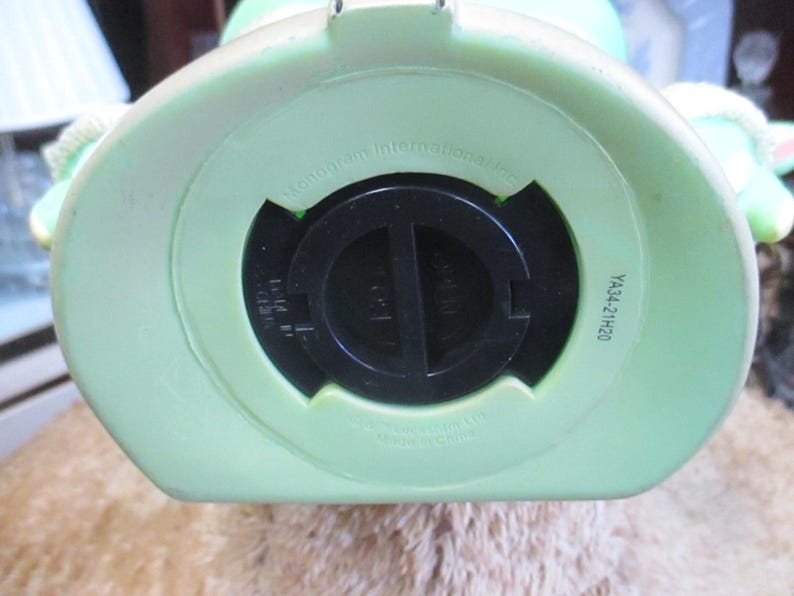 May include: Bottom view of a light green piggy bank with a black coin slot cover. The piggy bank has a round shape with a flat bottom. The text "YA34-21H20" is printed on the side.