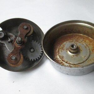 May include: A disassembled metal bicycle bell consisting of two circular components. One part features an internal gear mechanism with a lever, while the other is a hollow dome. Both pieces show visible signs of oxidation and rust on the metal.