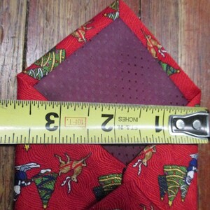 May include: Red Christmas-themed necktie with a festive pattern of reindeer, Christmas trees, and stars. The tie is measured with a tape measure, showing a width of approximately 2 inches.
