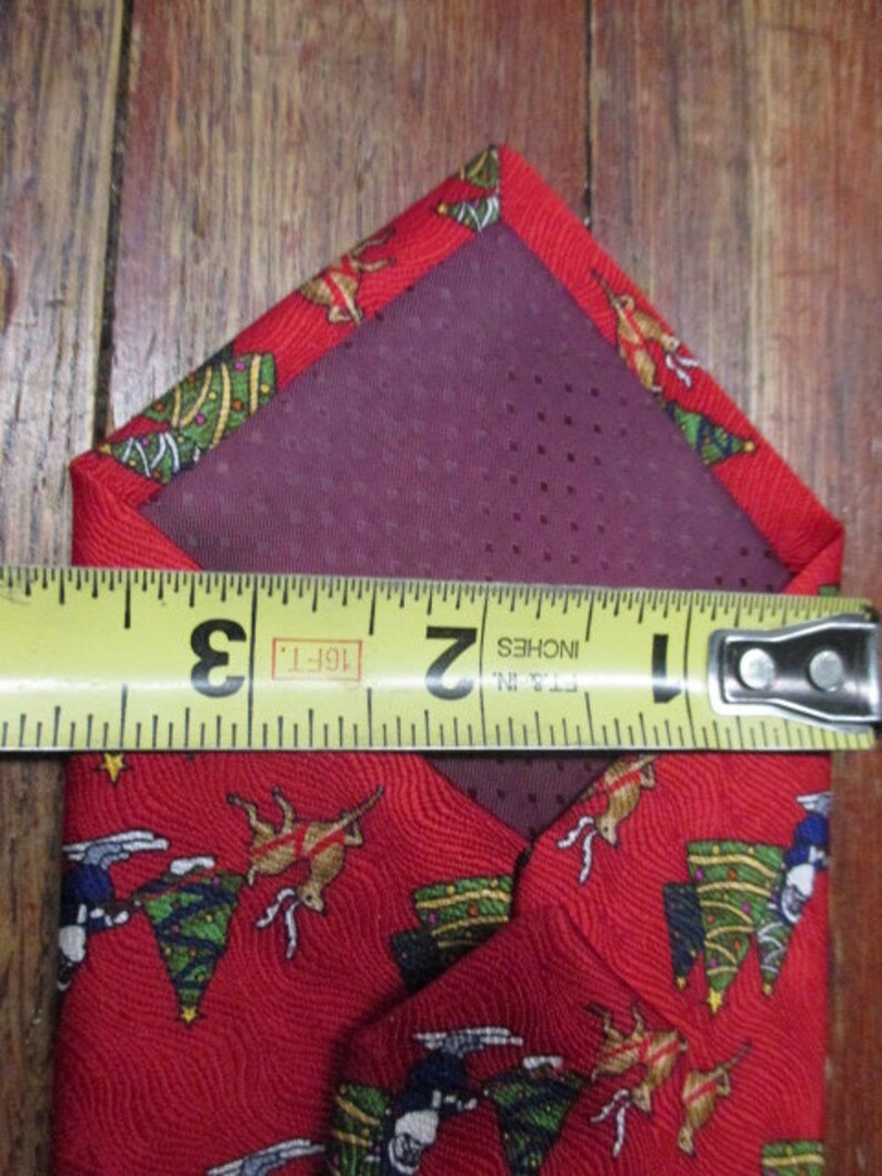 May include: Red Christmas-themed necktie with a festive pattern of reindeer, Christmas trees, and stars. The tie is measured with a tape measure, showing a width of approximately 2 inches.