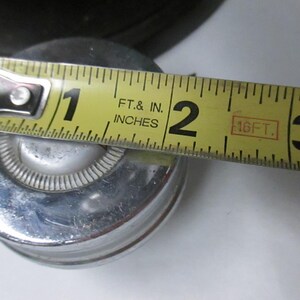May include: A circular, metallic object with a polished silver finish and a central white button-like detail. A yellow measuring tape is positioned across the top, indicating a diameter of approximately 2 inches for the circular component.