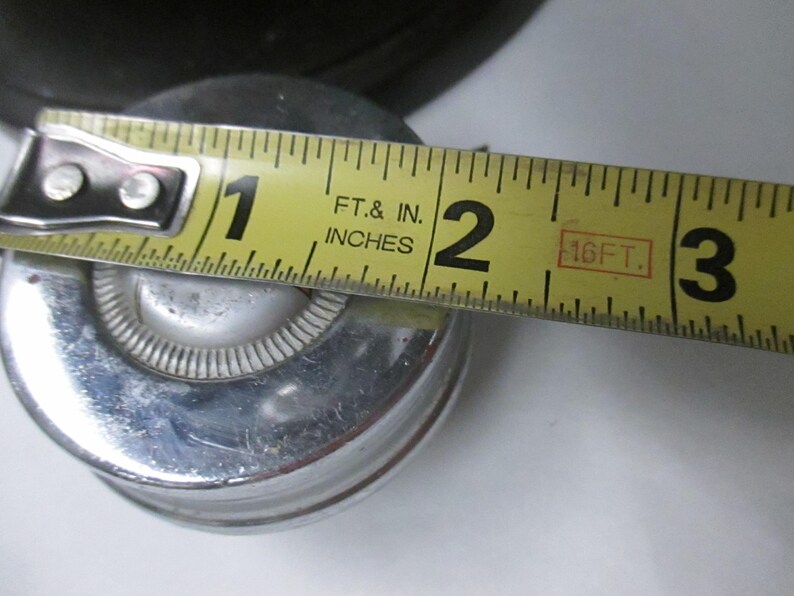 May include: A circular, metallic object with a polished silver finish and a central white button-like detail. A yellow measuring tape is positioned across the top, indicating a diameter of approximately 2 inches for the circular component.