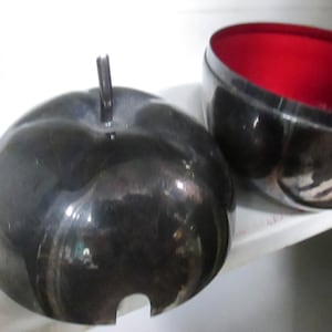 May include: A decorative, apple-shaped container with a dark, metallic finish. The lid features a stem-like handle. The interior of the accompanying bowl is a vibrant red. The container is designed to hold small items.