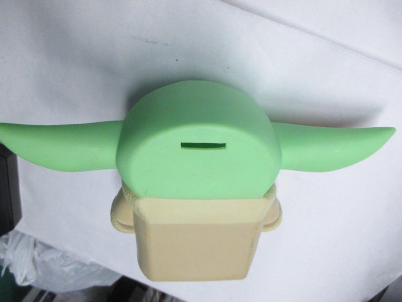 May include: A light green ceramic coin bank shaped like a character with large ears. The bank has a slot on top for inserting coins and a tan-colored base.