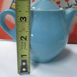 May include: A light blue ceramic teapot with a lid and handle. The teapot is approximately 3 inches tall. The teapot has a spout and a rounded body. The background is a red and white patterned design.