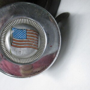 May include: A circular chrome metal knob featuring a central emblem with a graphic illustration of the American flag. The flag design displays a blue field with white stars and red and white horizontal stripes, surrounded by a textured silver border.