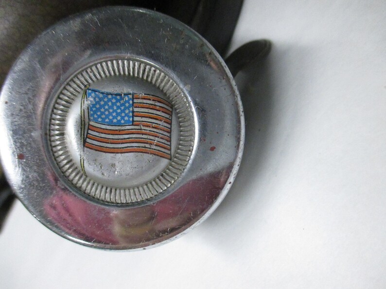 May include: A circular chrome metal knob featuring a central emblem with a graphic illustration of the American flag. The flag design displays a blue field with white stars and red and white horizontal stripes, surrounded by a textured silver border.