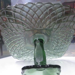 May include: A light green glass pedestal bowl featuring a raised lattice pattern on the underside of the bowl. The piece has a central fluted stem and a square-shaped base with scalloped edges, showcasing a translucent finish throughout the glass.