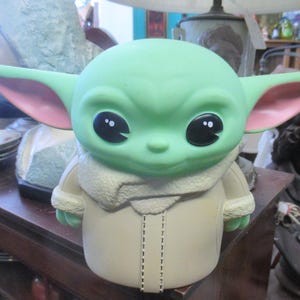 May include: A green figurine of a character with large black eyes, large ears with pink insides, and a tan coat. The figurine is a stylized representation of a popular character from a science fiction franchise.