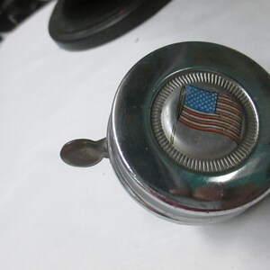 May include: A circular chrome bicycle bell featuring a central emblem with a graphic of the American flag. The bell has a polished metallic finish and a small lever extending from the side for activation, designed for mounting on handlebars.