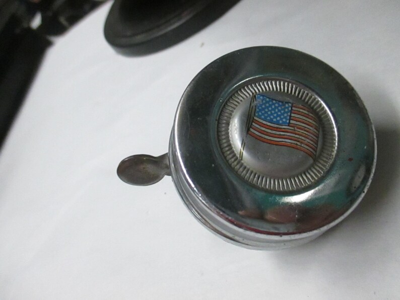 May include: A circular chrome bicycle bell featuring a central emblem with a graphic of the American flag. The bell has a polished metallic finish and a small lever extending from the side for activation, designed for mounting on handlebars.