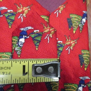 May include: Red Christmas tie with a festive pattern featuring reindeer, Christmas trees, and skiers. The tie is approximately 1 inch wide, as measured by a yellow tape measure.