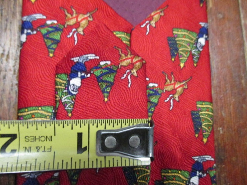 May include: Red Christmas tie with a festive pattern featuring reindeer, Christmas trees, and skiers. The tie is approximately 1 inch wide, as measured by a yellow tape measure.