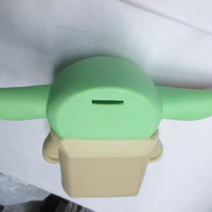 May include: A light green ceramic coin bank shaped like a character with large ears. The bank has a slot on top for inserting coins and a tan-colored base.