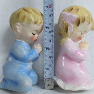 May include: Two ceramic figurines of children kneeling in prayer. The boy wears blue pajamas, and the girl wears a pink dress. Both have blonde hair and are approximately 3 inches tall. The figurines are vintage and have a glossy finish.