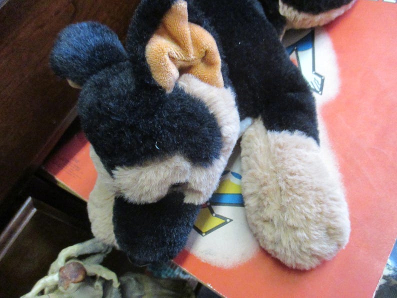 May include: A plush dog toy with black, tan, and white fur. The toy has floppy ears and a soft, cuddly appearance. The dog's paws are a light tan color. The toy is lying on a red surface.