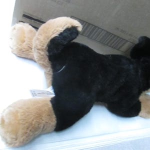 May include: A plush toy dog in a lying position, featuring black fur on its body and tan fur on its paws and ears. The toy is positioned on a white surface, with a cardboard box partially visible in the background.