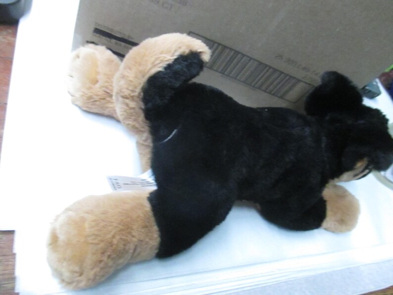 May include: A plush toy dog in a lying position, featuring black fur on its body and tan fur on its paws and ears. The toy is positioned on a white surface, with a cardboard box partially visible in the background.